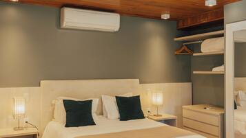 A bedroom with a wooden ceiling, a bed with white bedding, a bedside table, a nightstand, a wall-mounted air conditioner, and a wardrobe with a mirror.