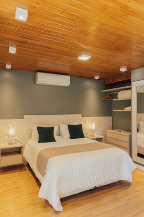 A bedroom with a wooden ceiling, a bed with white bedding, a bedside table, a nightstand, a wall-mounted air conditioner, and a wardrobe with a mirror.