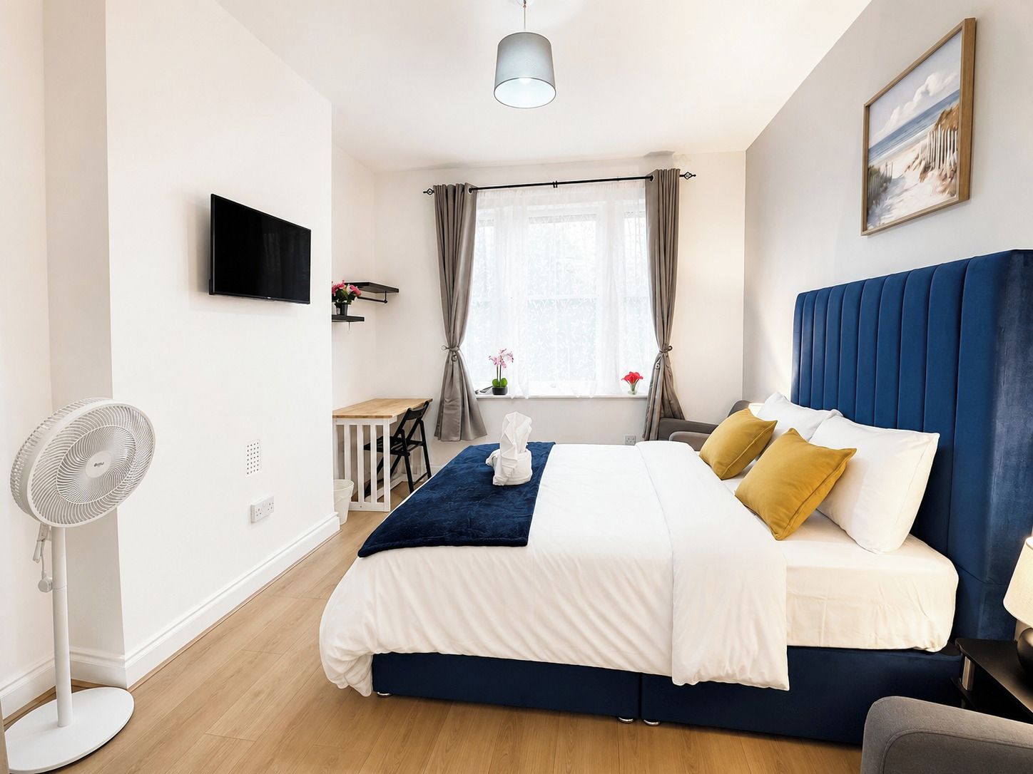 A bedroom with a bed, a desk, a chair, a standing fan, and a painting on the wall.