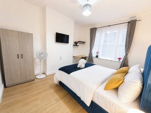 Um quarto com cama, guarda-roupa, televisão, janela com cortinas e ventilador.