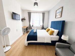 Ein Hotelzimmer mit Bett, Nachttisch, Lampe, Stuhl, Schreibtisch und einem an der Wand befestigten Fernseher.