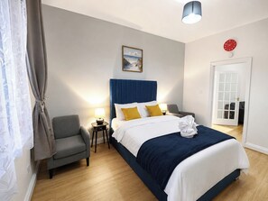 Ein Hotelzimmer mit einem Bett, einem Sessel, einem Nachttisch mit Lampe, einem Bild an der Wand und einer Tür.