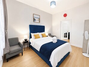 Ein Hotelzimmer mit Bett, Sessel, Nachttisch, Lampe, Uhr und Tür.