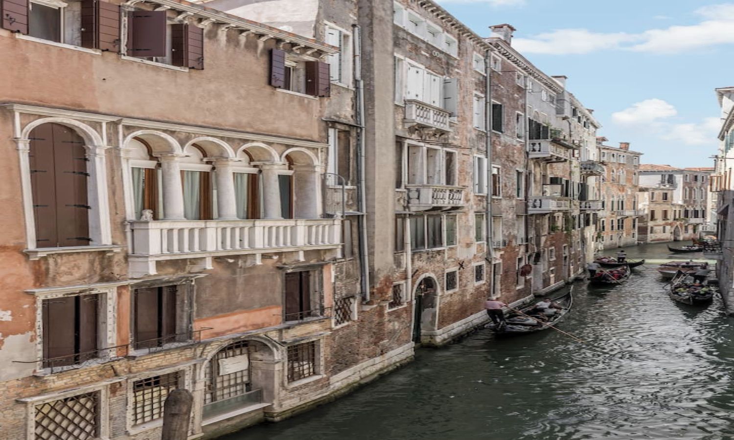 A narrow canal flanked by historic buildings with arched windows and balconies, a gondola in the water, and a person rowing a boat.