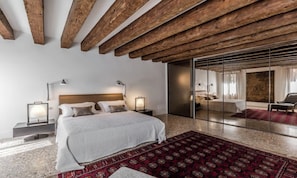 Quarto moderno com uma cama grande, mesas de cabeceira, um tapete estampado em vermelho e teto de madeira.