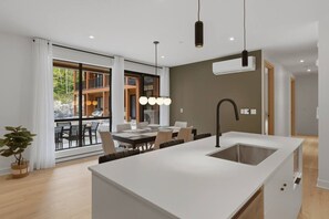 A modern kitchen with a central island, a dining area with a table and chairs, and a view through large windows. - THE VALO Ostrya 3BDR Mont-Tremblant (Mont-Tremblant)