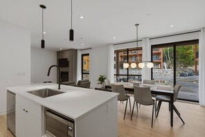 A modern kitchen with a central island, dining area with chairs, and large windows offering a view of the outside. - THE VALO Ostrya 3BDR Mont-Tremblant (Mont-Tremblant)