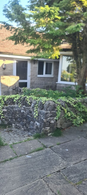 A stone wall with greenery, a paved area, and a building with windows and a door.