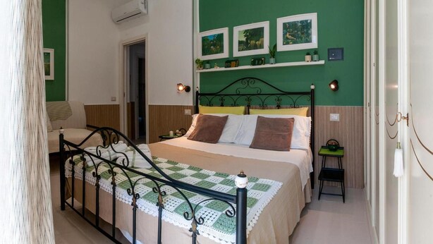 Une chambre à coucher avec un lit, une table de chevet, une chaise, un mur vert orné de tableaux et une porte.