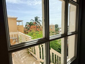 A view through a window with a white railing, a balcony, and a garden with palm trees and flowering plants. - 1-Bedroom Apartment in Pelican Key with Shared Pool & Balcony (Koolbaai)