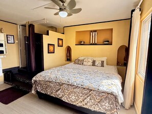 A bedroom with a bed, a ceiling fan, a shelf with decorative items, and a door with a curtain. - 1-Bedroom Apartment in Pelican Key with Shared Pool & Balcony (Koolbaai)