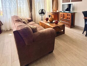 A living room with a sofa, a wooden coffee table, a fan, and a TV. - 1-Bedroom Apartment in Pelican Key with Shared Pool & Balcony (Koolbaai)