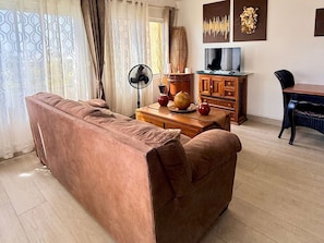 A living room with a sofa, a wooden coffee table, a TV, and a dining area with a chair. - 1-Bedroom Apartment in Pelican Key with Shared Pool & Balcony (Koolbaai)