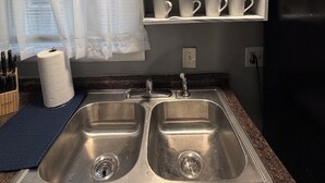 Una cocina con dos lavamanos de acero inoxidable, un dispensador de toallas de papel, un bloque para cuchillos y un estante con copas y tazas.