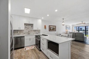 A modern kitchen with white cabinetry, a central island, and a dining area with a sofa and table. - Runaway Bay 2BR|Steps to Beach + Heated pool/gym (Bradenton Beach)
