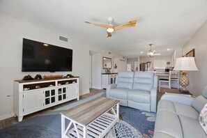 A living room with a television, a white cabinet, a blue sofa, a wooden coffee table, and a ceiling fan. - Runaway Bay 2BR|Steps to Beach + Heated pool/gym (Bradenton Beach)
