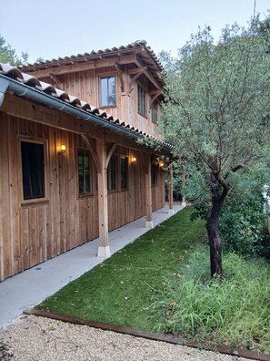 A wooden building with a covered porch, a well-maintained lawn, and a tree. - Lachanau Cabin: Relaxation and Fun with a Covered Terrace and Walkways Near the Lake (Hourtin)