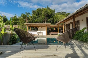 Ein Poolbereich mit zwei Korbstühlen, einer Holzterrasse und einer überdachten Sitzgelegenheit mit Tisch und Stühlen.
