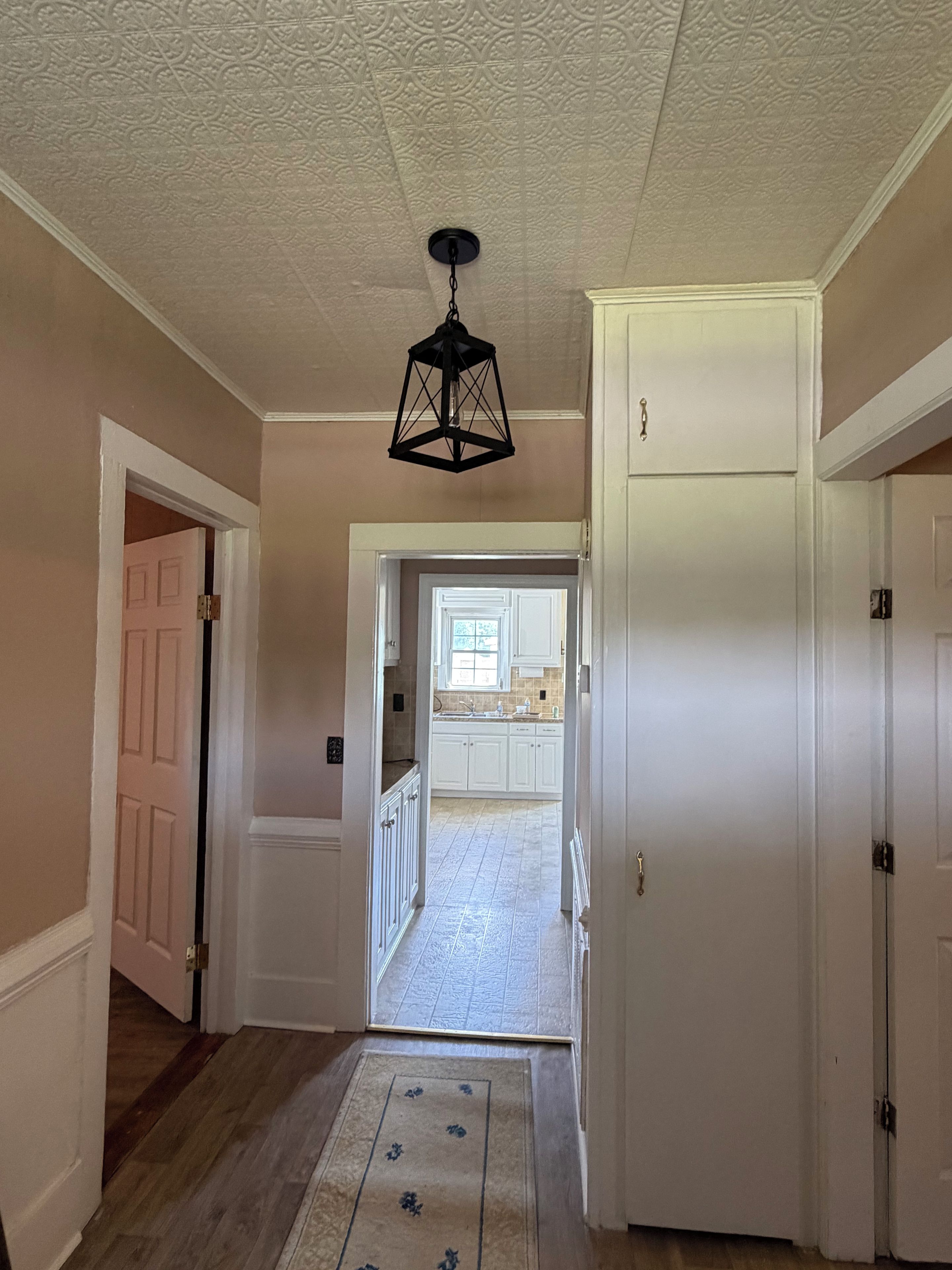 A hallway with a patterned ceiling, a black hanging light fixture, and a view into a kitchen area with white cabinets and a window.