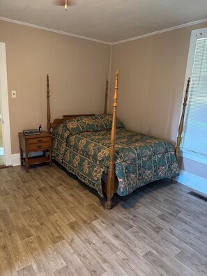 Un dormitorio con cama con dosel, mesita de noche con cajones, suelo de madera y ventana con persianas.