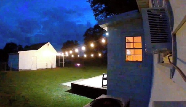 Un patio trasero con un porche cubierto, un edificio blanco y luces colgantes.