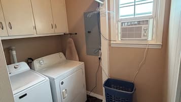 A laundry room with two white washers and dryers, a blue laundry basket, and a window with an air conditioning unit.