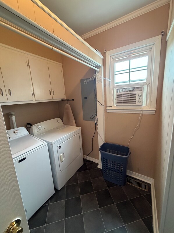 A laundry room with two white washers and dryers, a blue laundry basket, and a window with an air conditioning unit.