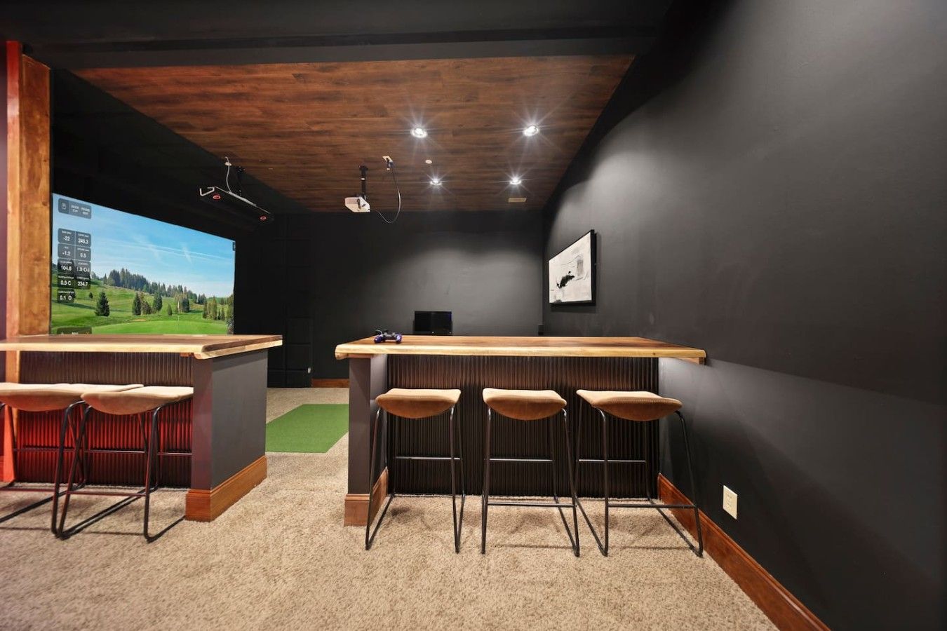 A bar area with a wooden counter, bar stools, and a television screen displaying a landscape.