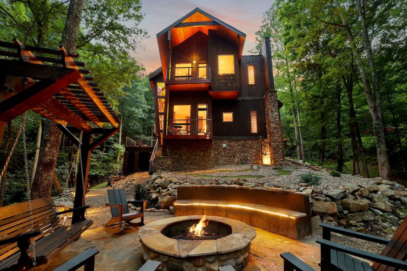 Een meerlaagse cabin met een stenen fundering, omgeven door een bos. Er is een vuurkuil met zitplaatsen en een houten terras.