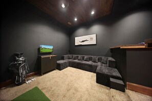 Ein Zimmer mit einer Eckcouch, einem Fernseher, einer Golfbag und einem gerahmten Bild an der Wand.
