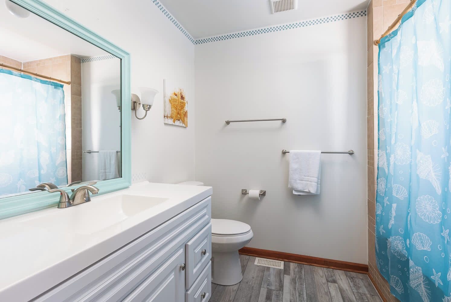 Une salle de bain avec un meuble-lavabo blanc, un grand miroir, des toilettes et un rideau de douche orné d’un motif sur le thème de la mer.