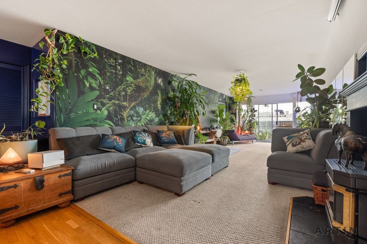 A modern living room with a sectional sofa, a coffee table, and a fireplace. There are multiple plants and a large wall mural.