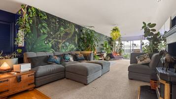 A modern living room with a sectional sofa, a coffee table, and a fireplace. There are multiple plants and a large wall mural.