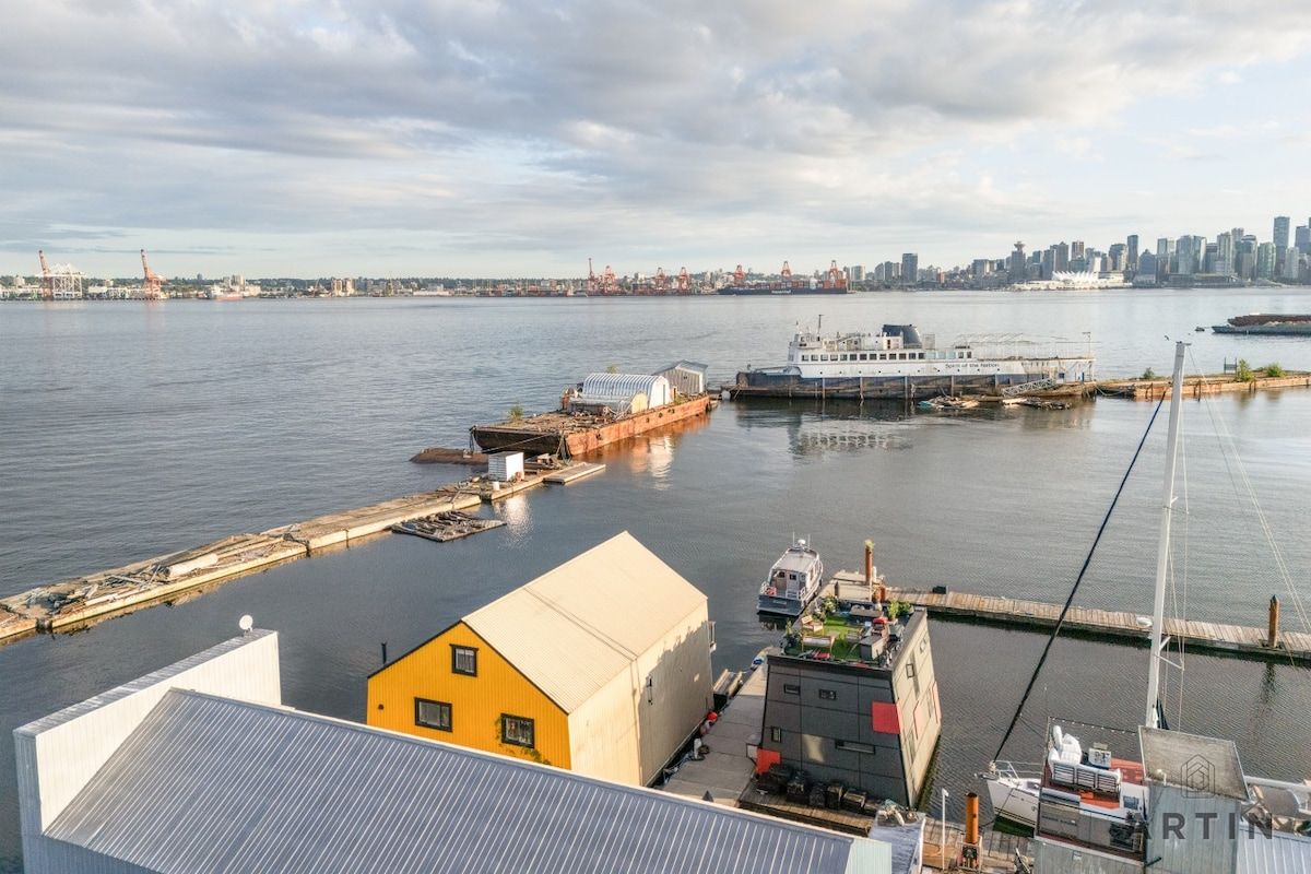 A waterfront view with a large white boat docked at a pier, a yellow building, and a city skyline in the distance.