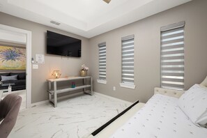 Un dormitorio con una cama, un televisor montado en la pared, una mesita consola y una ventana con persianas.