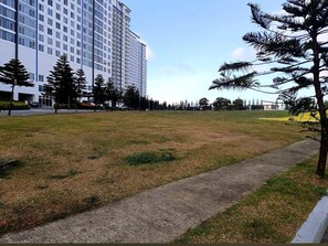 A grassy area with a concrete path, a tall building in the background, and several trees. - Cozy 1-Bedroom Tagaytay Condo with Balcony & Wi-Fi Near Taal View and Sky Ranch (Tagaytay City)