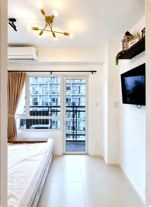 A modern bedroom with a bed, a TV mounted on the wall, a ceiling light fixture, and a window with curtains. - Cozy 1-Bedroom Tagaytay Condo with Balcony & Wi-Fi Near Taal View and Sky Ranch (Tagaytay City)
