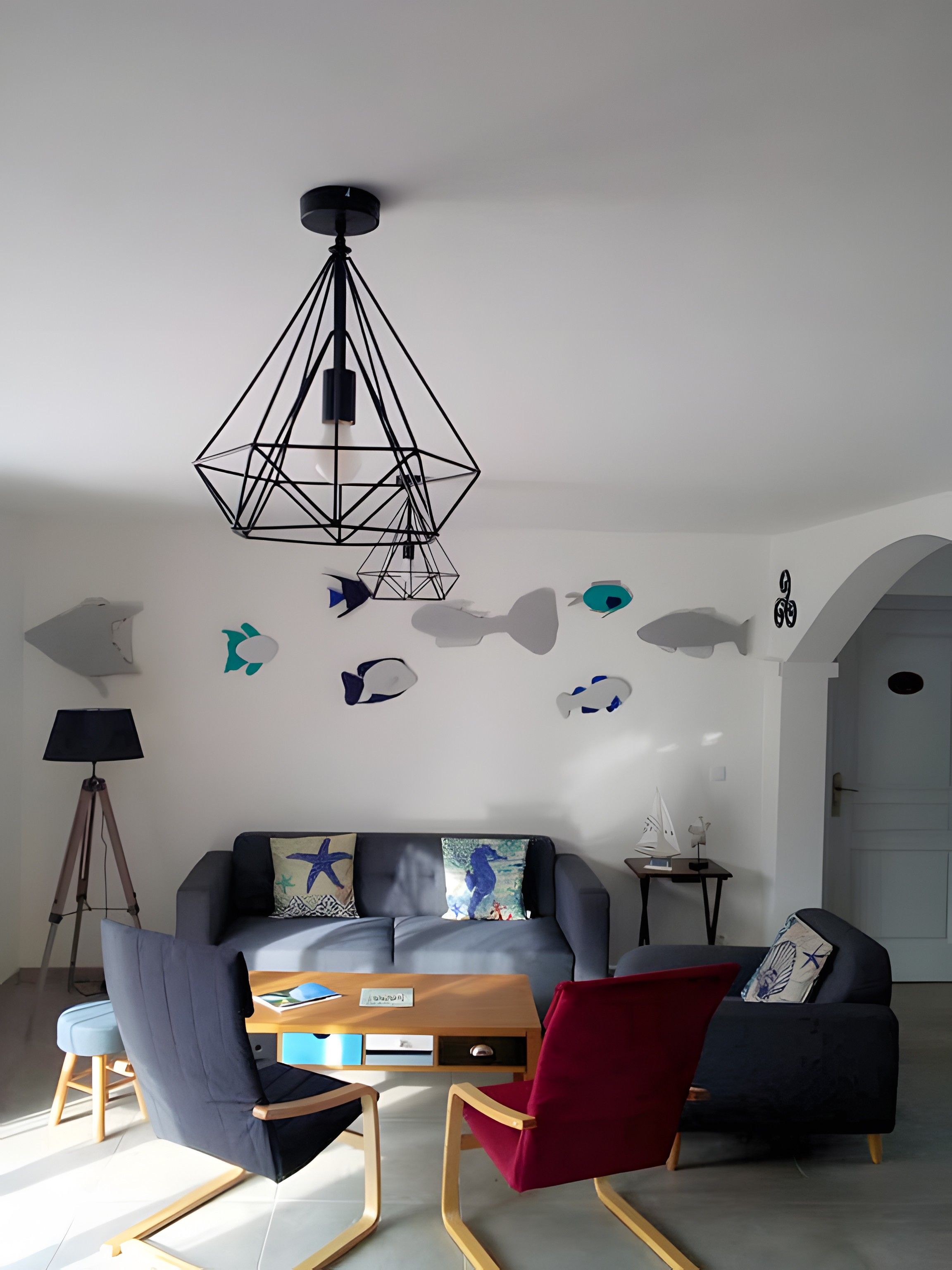 A modern living room with a geometric ceiling light, a sofa with patterned cushions, a red chair, and a small table with a lamp.