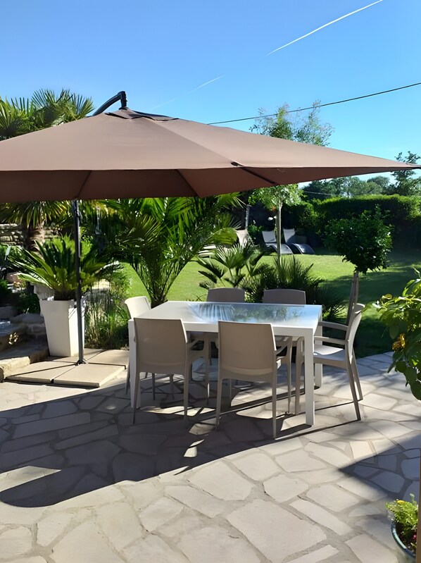 An outdoor patio with a table and chairs under a large umbrella, surrounded by greenery.
