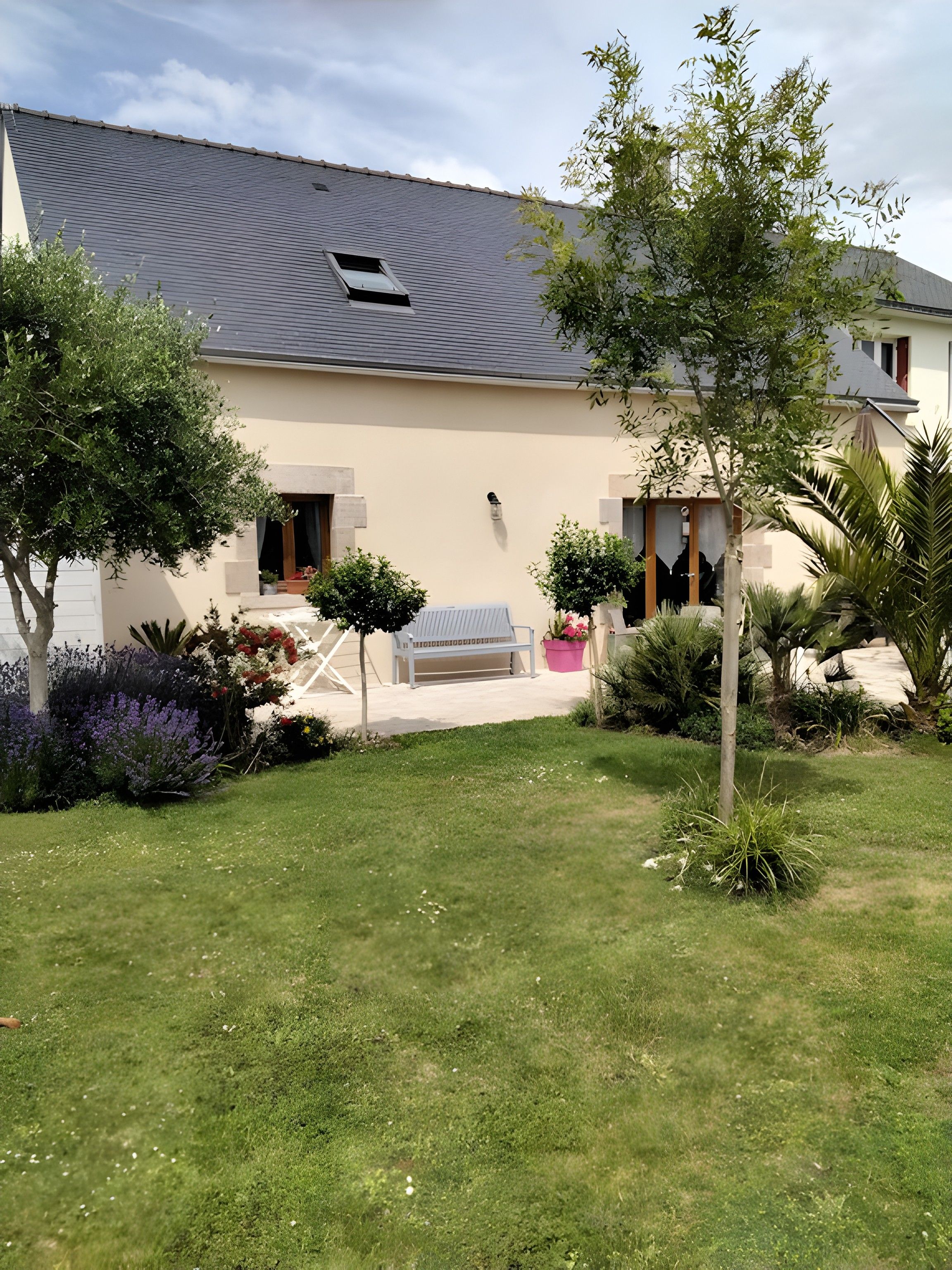 Une maison avec un toit en pente, un petit porche doté d’un banc et un jardin bien entretenu, agrémenté de diverses plantes et fleurs.