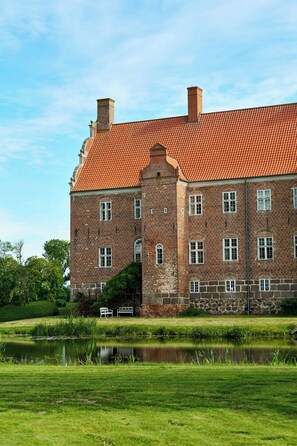 Un grand bâtiment historique, doté d’un toit recouvert de tuiles rouges et de murs en briques, situé à côté d’un étang paisible, avec une pelouse en contrebas.