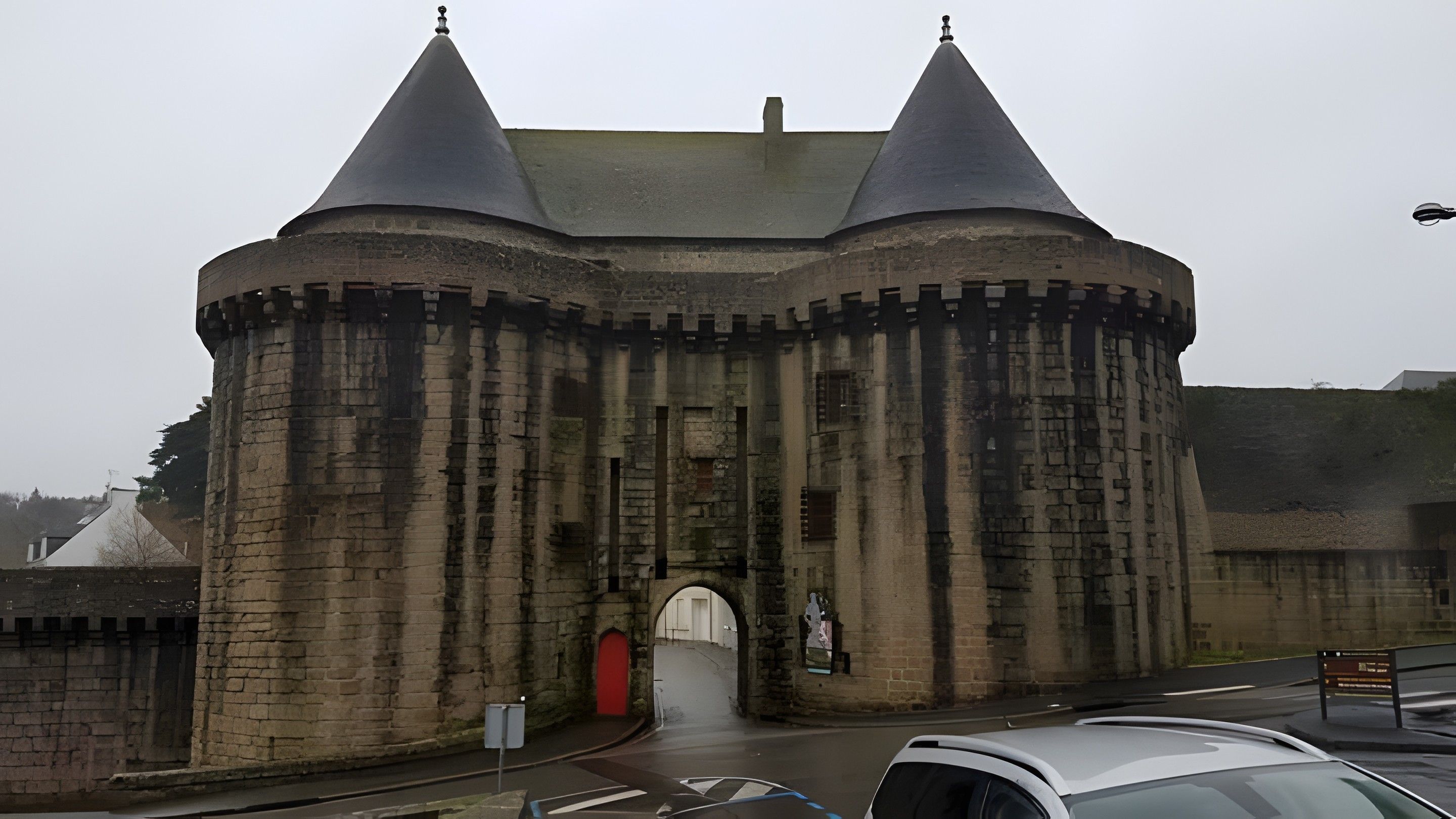 Een historisch gebouw van natuursteen met twee kegelvormige torens, een centrale boogvormige ingang en een rode deur.