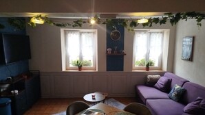 A living room with a purple sofa, a dining table with placemats, and two windows with plants. - The Great Cozy Safari (Petersbach)