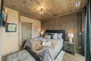 Un dormitorio con techo de madera, una cama con muchos almohadones, una mesita de noche con lámpara y un cuadro enmarcado en la pared.