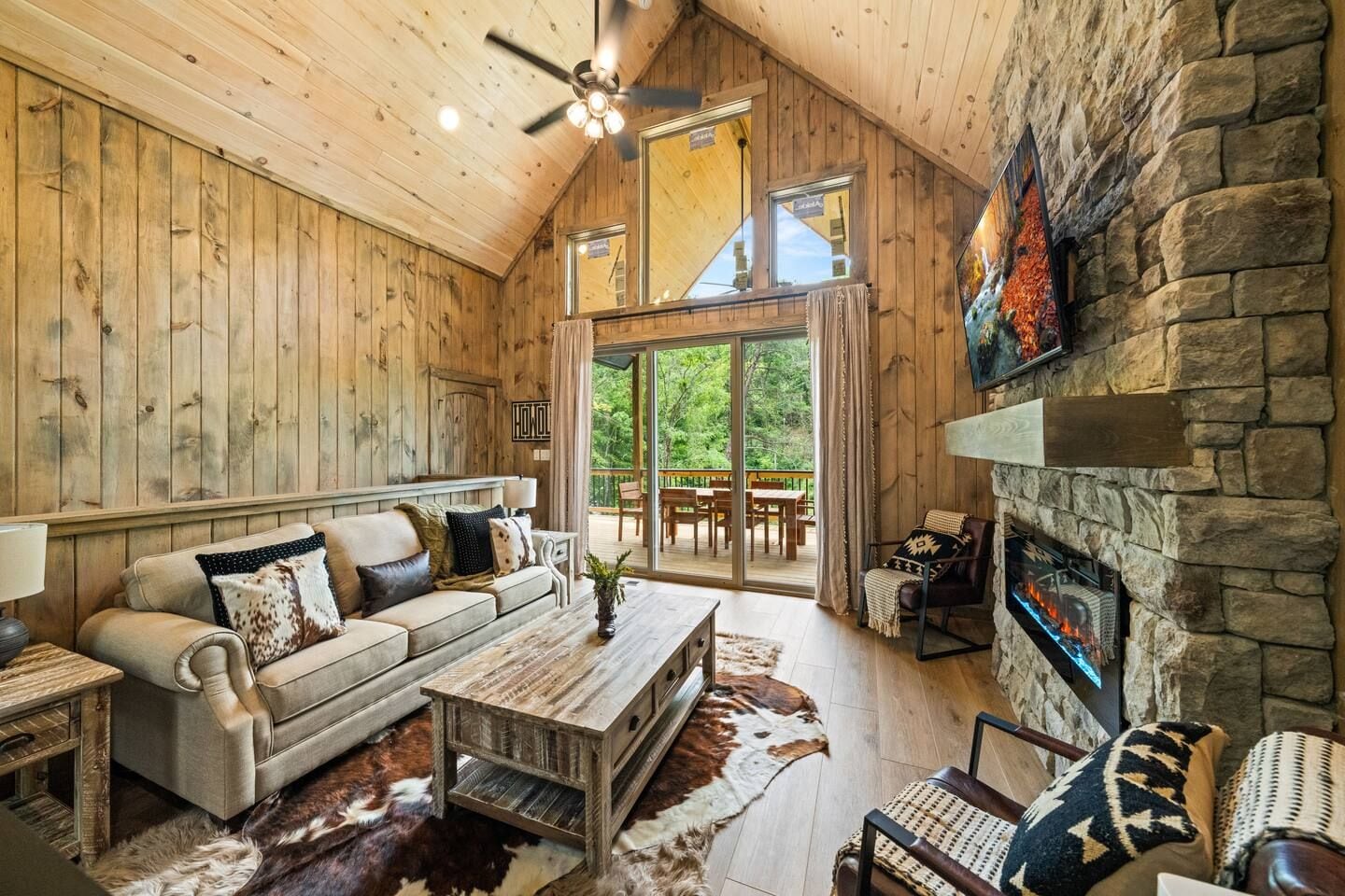 A cozy cabin interior with a stone fireplace, wooden walls, a sofa, and a dining area with a view of the outdoors.
