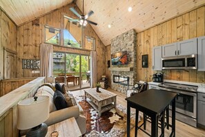 Interior rústico de una cabaña con detalles de madera, una chimenea de piedra, una cocina con electrodomésticos modernos y un gran ventanal corredizo que da a una terraza exterior.