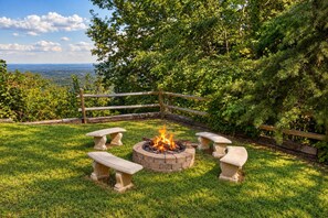 A fire pit with four benches in a grassy outdoor area with a wooden fence and trees in the background. - Radiant Sky Views | Hot Tub, Game Room, MORE! (Sevierville)