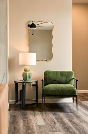 Ein Zimmer mit einem grünen Polsterstuhl, einem kleinen runden Tisch mit Lampe sowie einem an der Wand befestigten Spiegel mit goldener Rahmung.