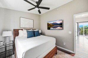 Ein Schlafzimmer mit Bett, Deckenventilator, Nachttisch, Lampe, einem Gemälde und Blick ins Freie.