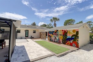 A residential property with a patio, a small lawn, and a colorful mural on the side of a building. - Cozy Sarasota Bungalow + Tropical Outdoor Space (Sarasota)